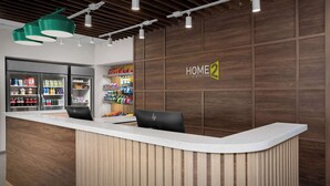 Et hotelreceptionsområde med en træreception, en digital skærm og et vægmonteret skilt med teksten HOME 2 SUITES BY HILTON.