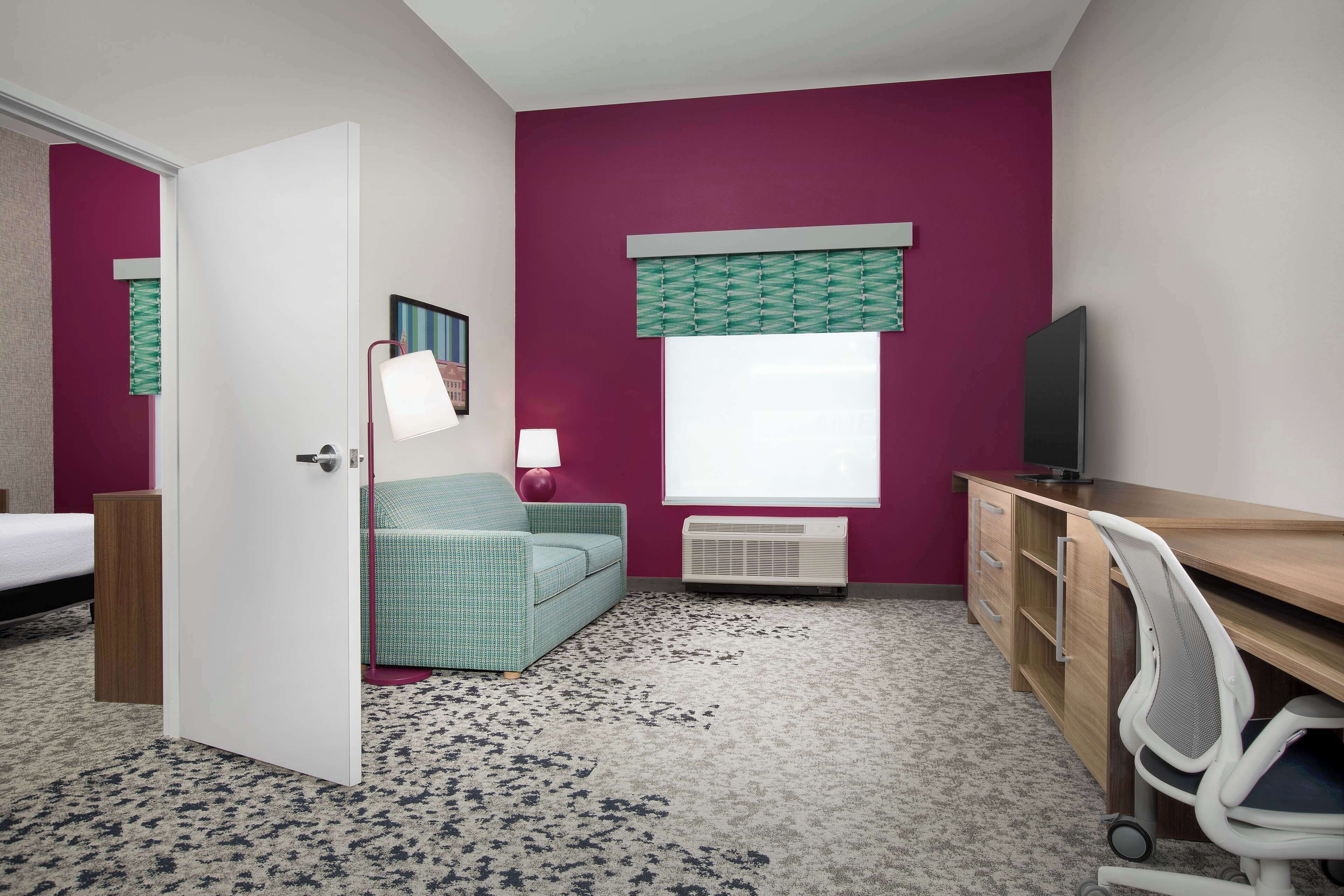 Quarto com cama, escrivaninha, cadeira, luminária, televisão e janela com cortina estampada.