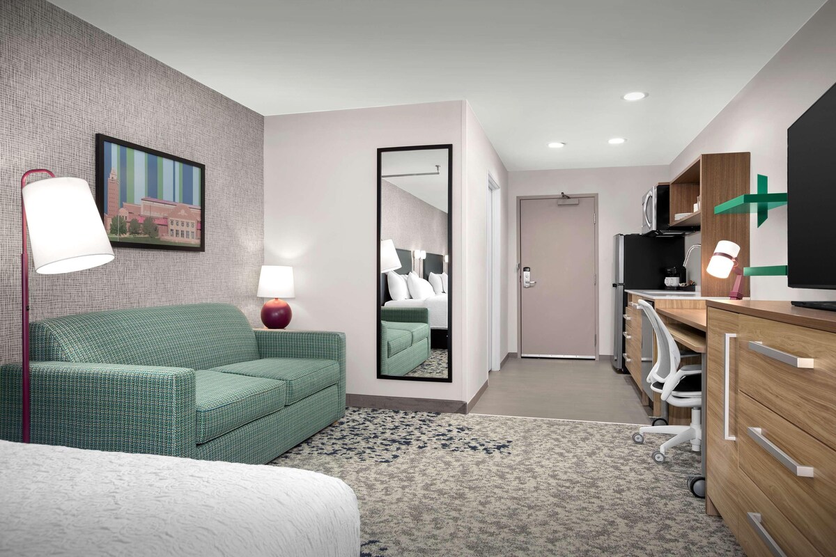 A modern hotel room with a green sofa, a large mirror, a flat-screen TV, and a desk with a computer.
