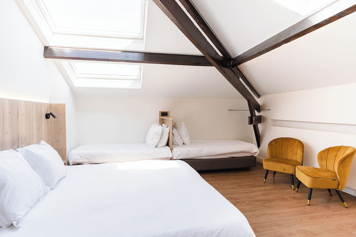 A bedroom with a sloped ceiling, wooden beams, a bed, two chairs, and a small bedside table.