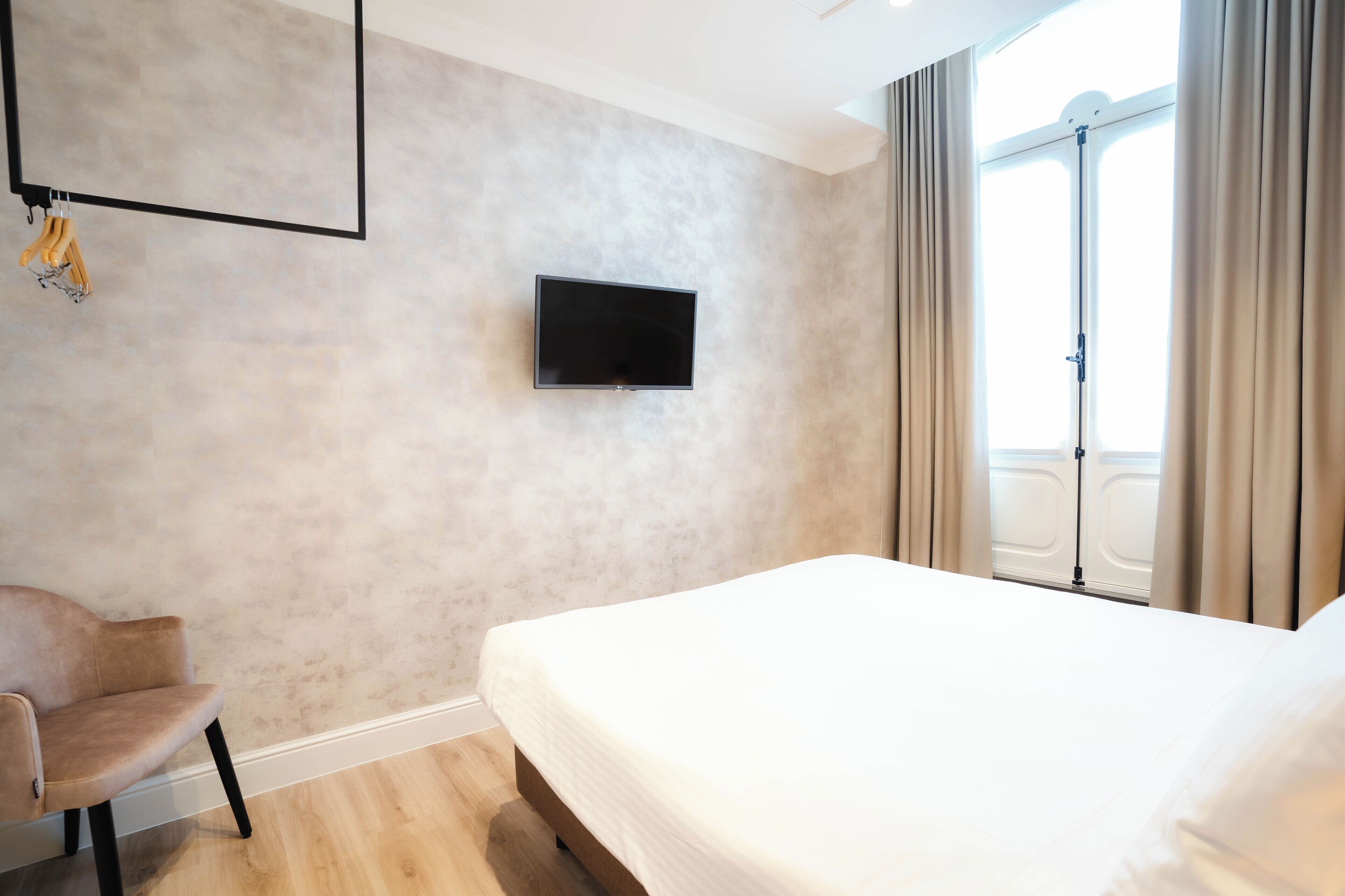 Una habitación de hotel con cama, televisor montado en la pared, una silla y una ventana con cortinas.