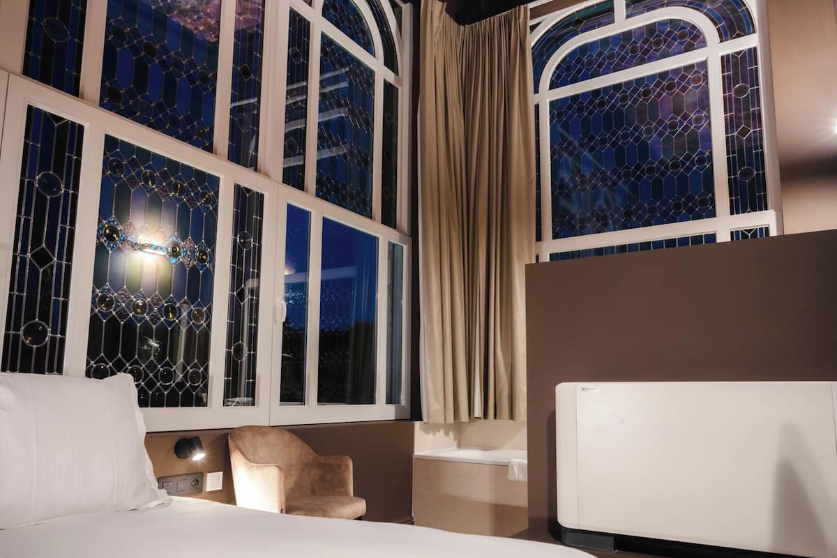 A hotel room with a large bed, a nightstand, a chair, a large window with stained glass, and a ceiling mural.