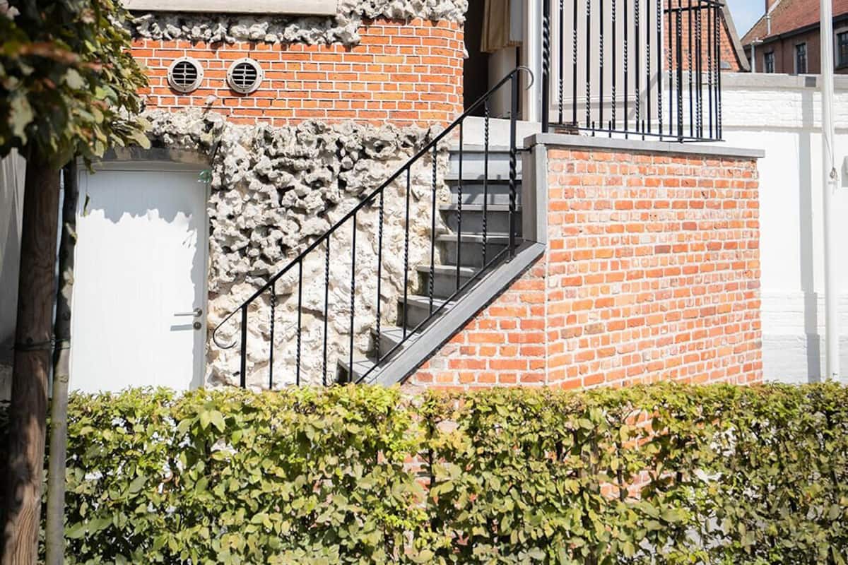 A brick house with a white door, a small balcony with black railings, and a stone wall.