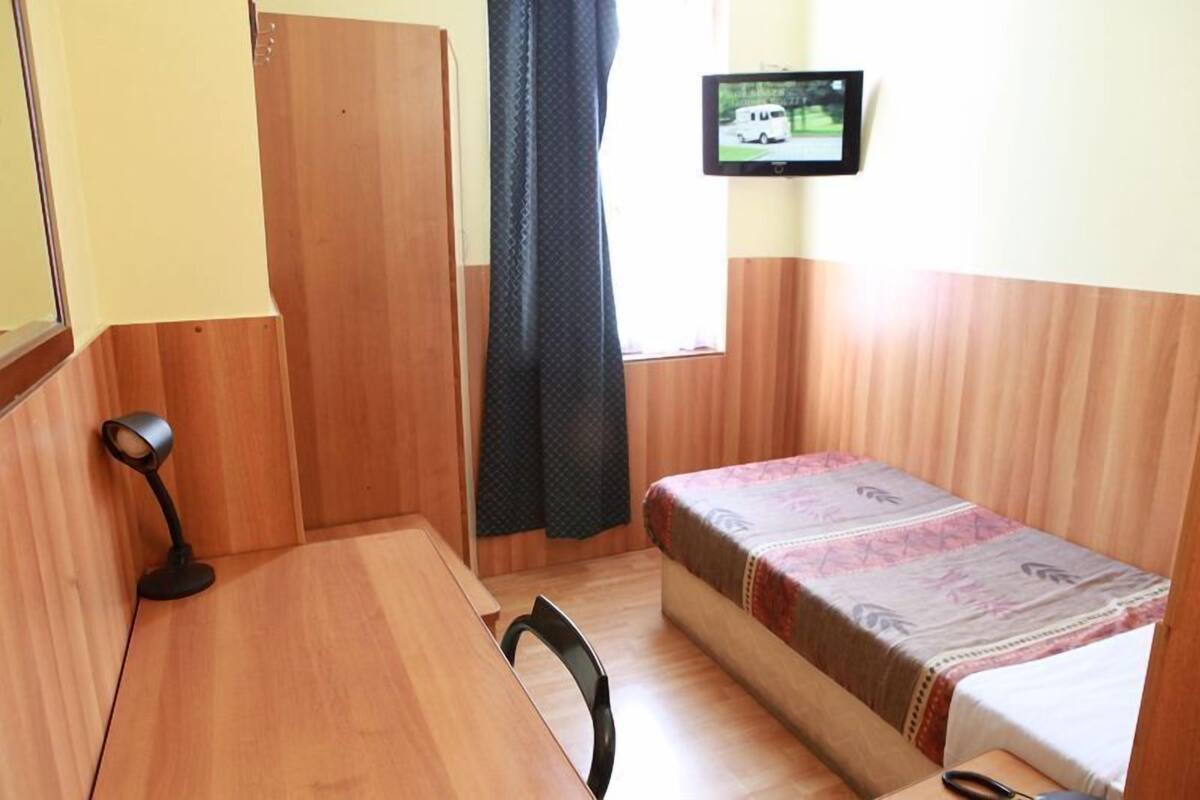 A small room with a single bed, a desk, a chair, a TV on the wall, and a window with curtains.