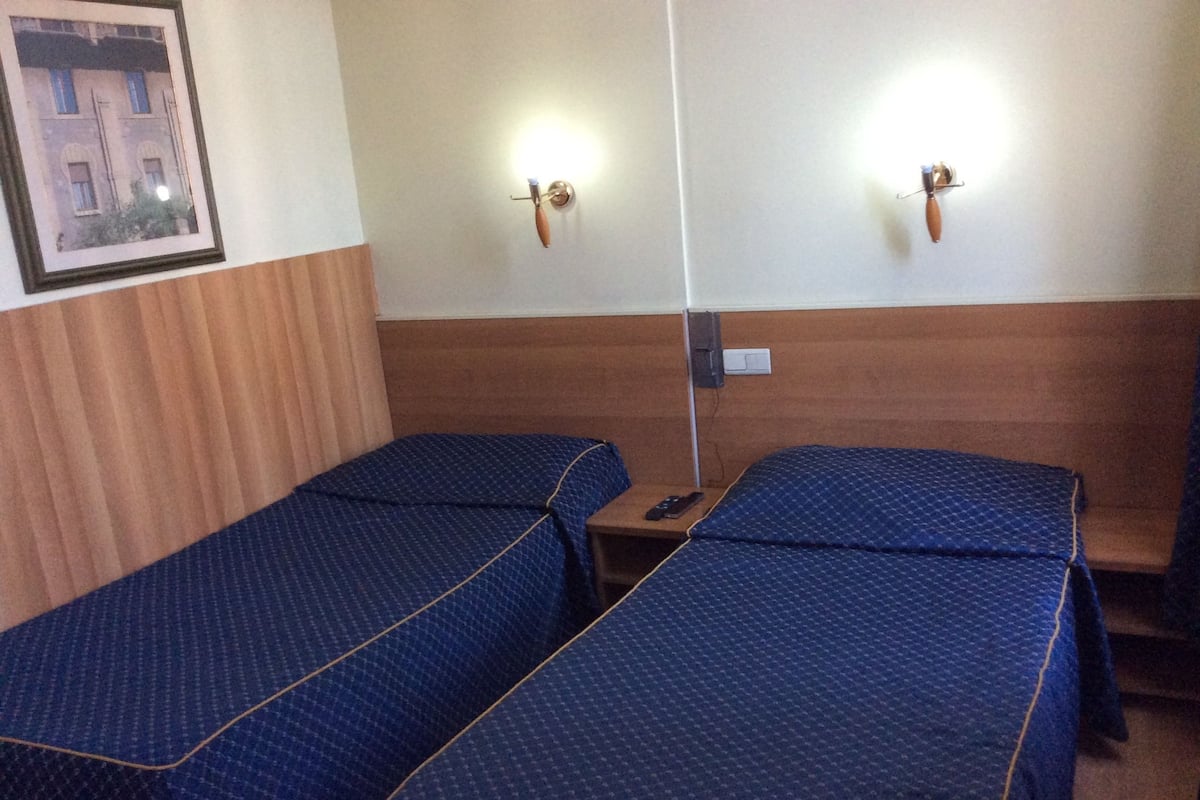 A hotel room with two single beds, wooden headboards, a small table, and a wall-mounted light fixture.