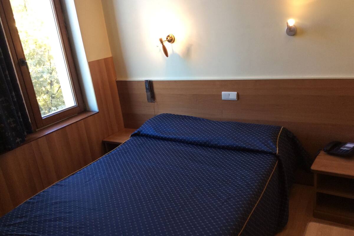 A single bed with a blue patterned bedspread in a room with wooden paneling, a bedside table with a phone, and a wall-mounted light fixture.