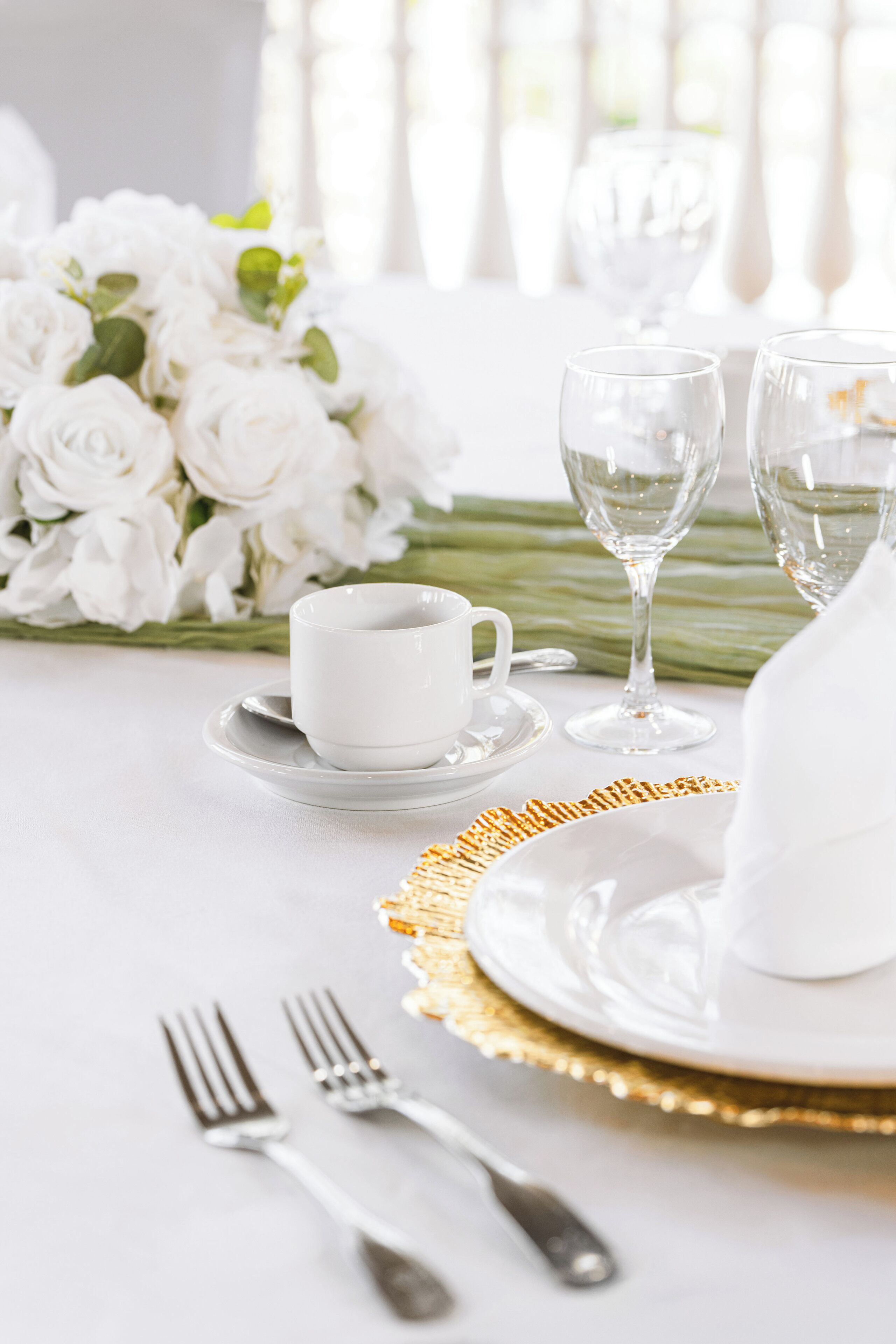 A table set with white dinnerware, including a cup, saucer, and glassware, with a gold-rimmed plate and a white napkin.