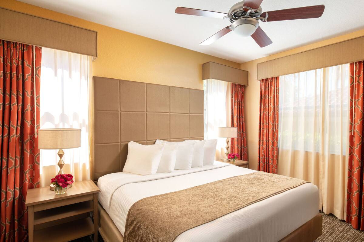 Una habitación de hotel con una cama grande, dos mesitas de noche, un ventilador de techo y cortinas rojas con estampado.