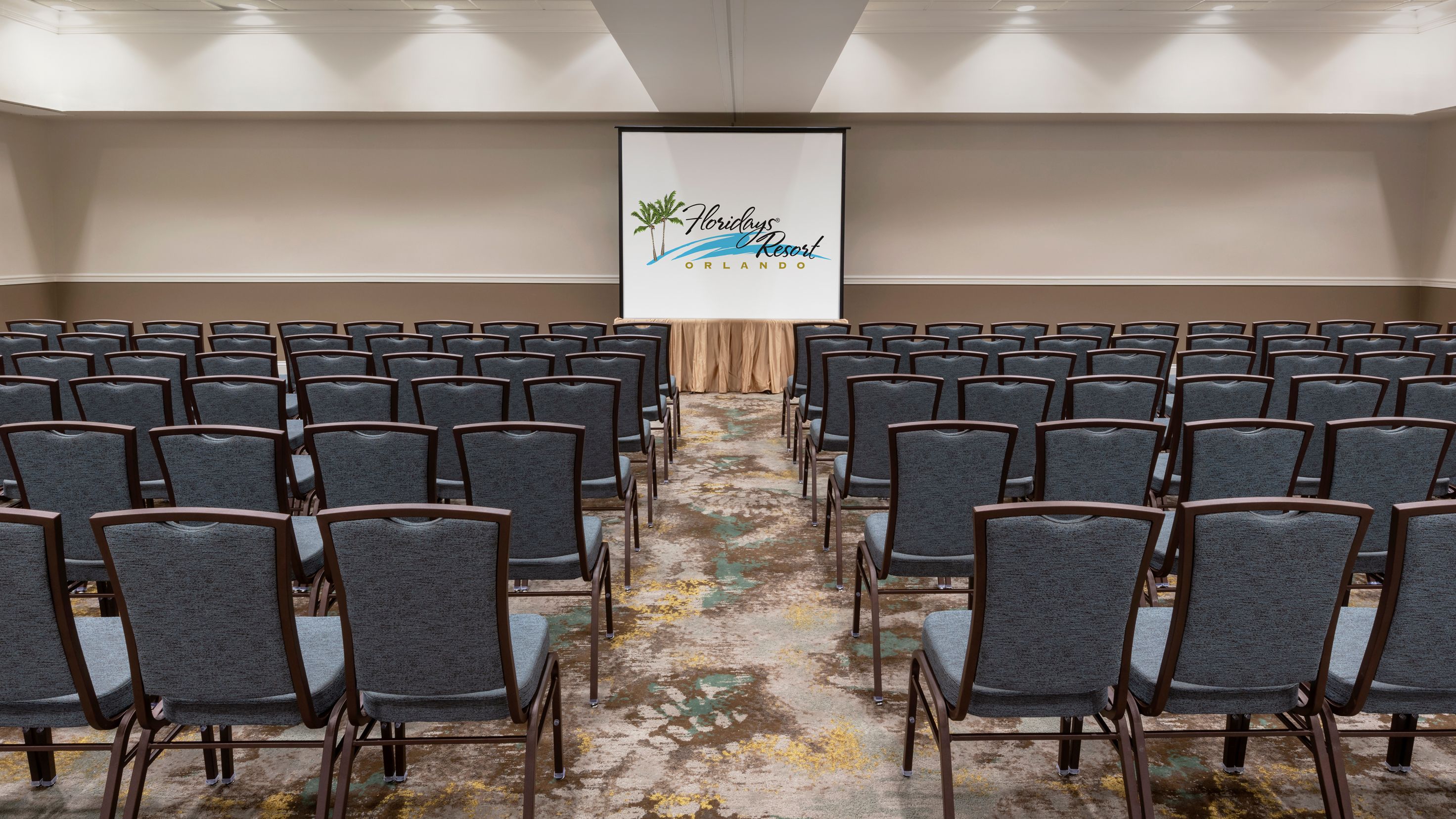 Une salle de conférence avec des rangées de chaises grises, un sol recouvert de moquette et un écran affichant le logo de « Florida's Resort Orlando ».