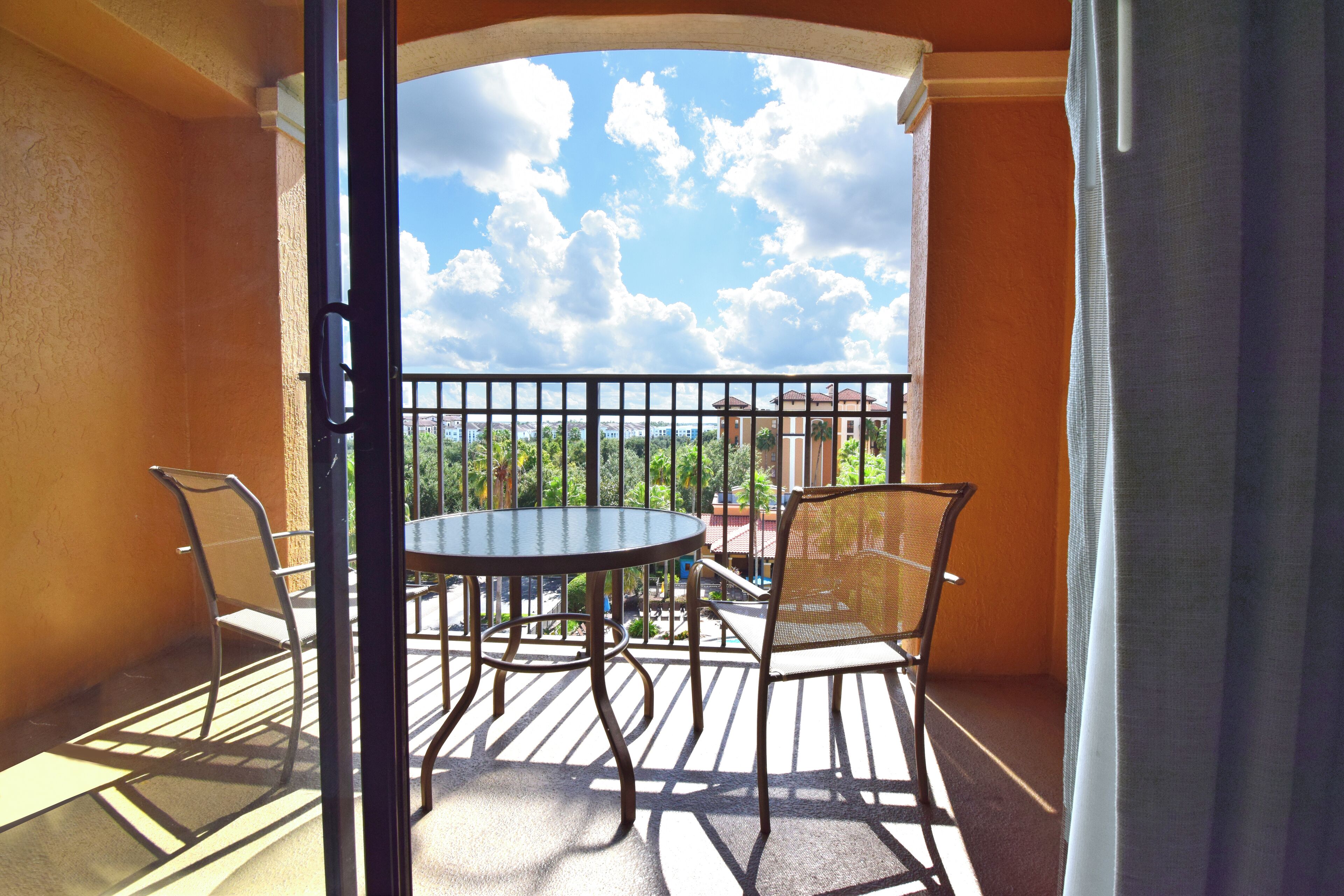 A balcony with a round table and two chairs, a view of greenery and buildings, and a clear sky.