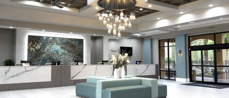 A modern reception area with a marble reception desk, a large abstract wall art piece, a seating area with a vase of flowers, and a ceiling with recessed lighting and a chandelier.