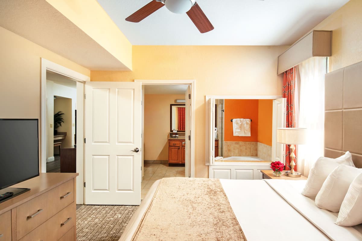 Habitación de hotel con cama, escritorio, televisor, armario y baño visible a través de una puerta abierta.