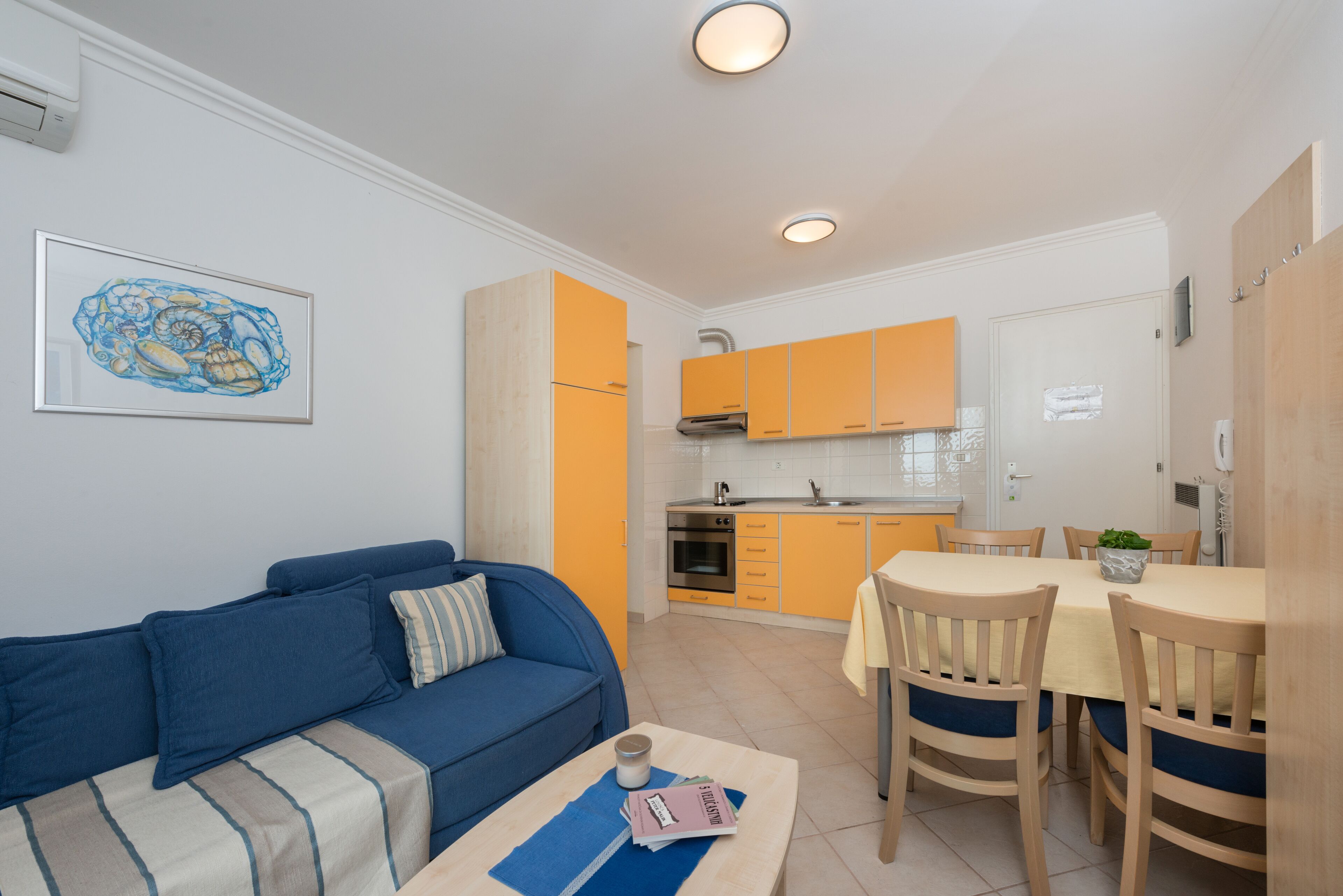 A compact living space with a blue sofa, a dining table with chairs, and a kitchen area with orange cabinets.