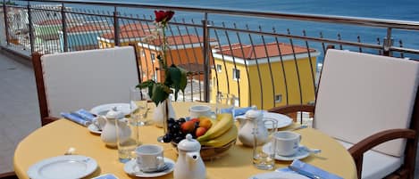 A table set for breakfast with a view of the sea, a rose, and a bowl of fruit.