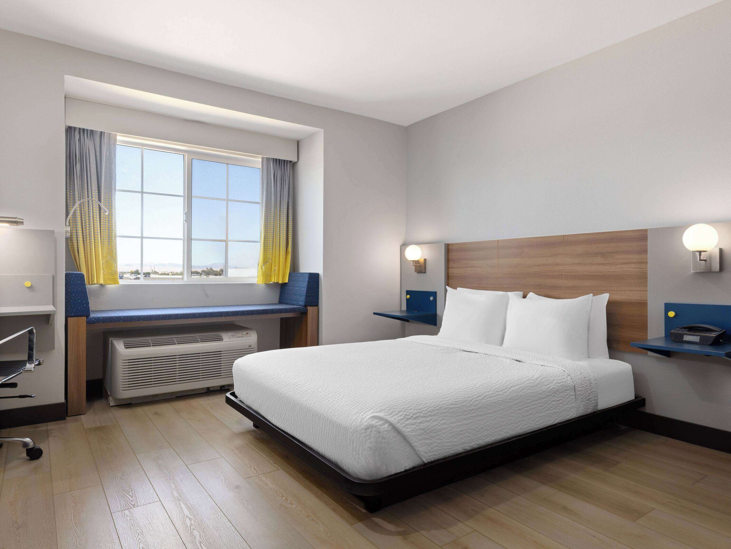 Habitación de hotel con cama, escritorio, silla, ventana con cortinas y lámpara de pared.