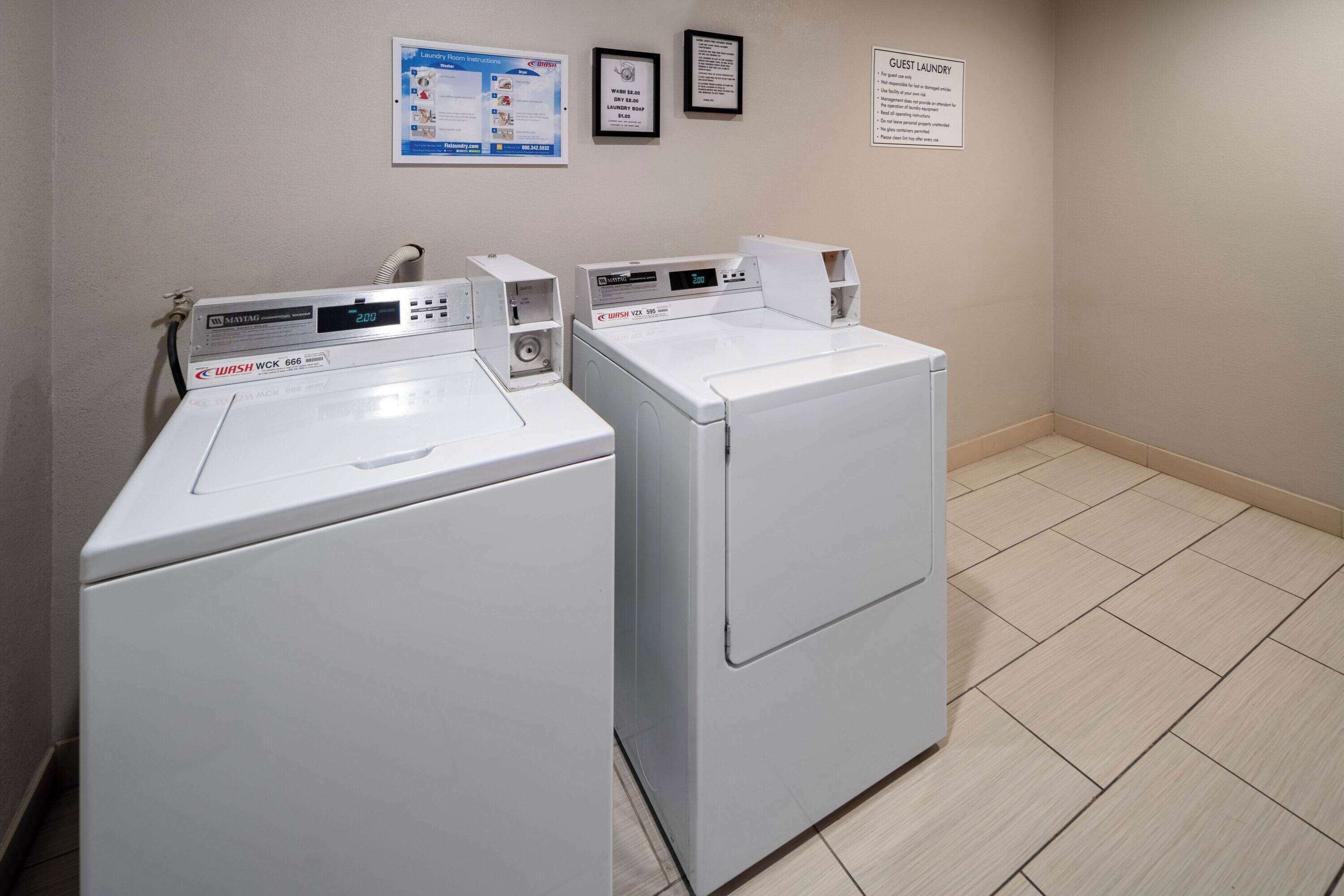 Un espace buanderie comprenant deux machines à laver et deux sèche-linge à chargement frontal, tous deux de couleur blanche, un panneau mural affichant des instructions et un avis encadré.