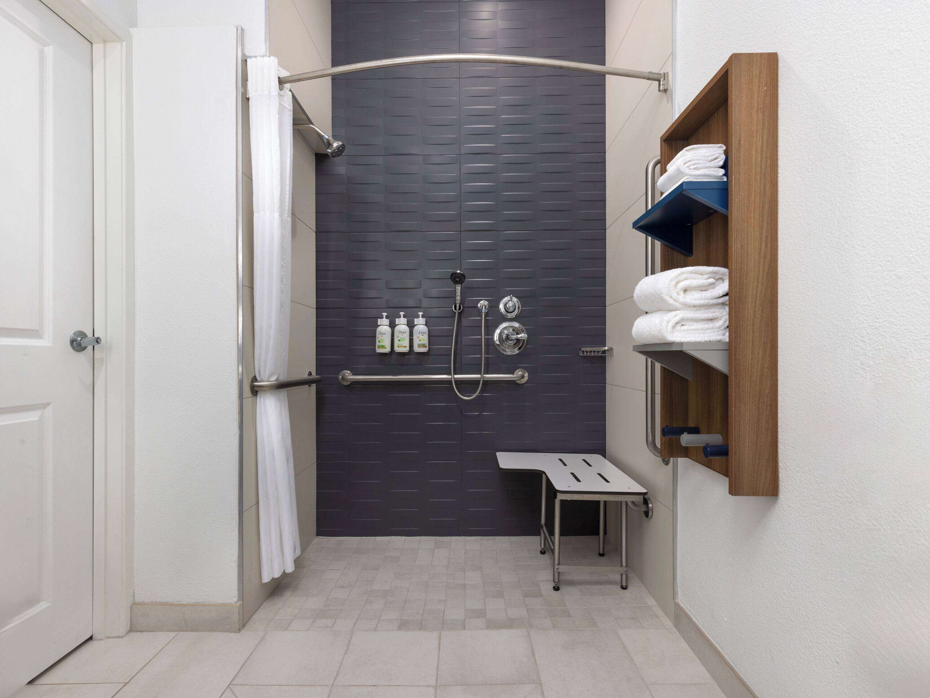 A narrow shower area with a dark tiled wall, a white door, a towel rack with folded towels, and a small table.
