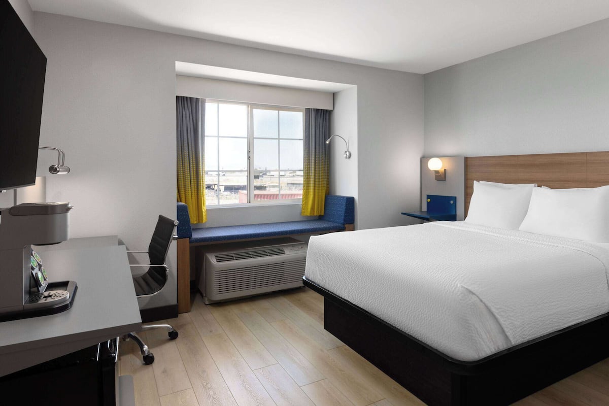 A hotel room with a bed, desk, chair, air conditioning unit, and a window with curtains.