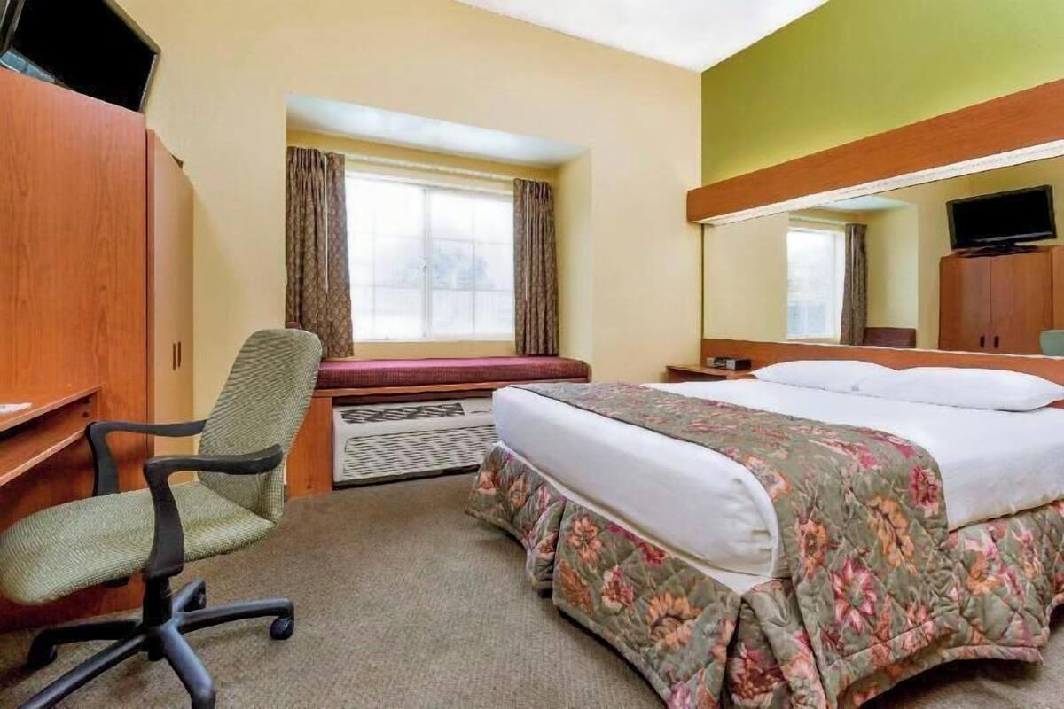 A hotel room with a bed, a desk with a chair, a television, and a window with curtains.