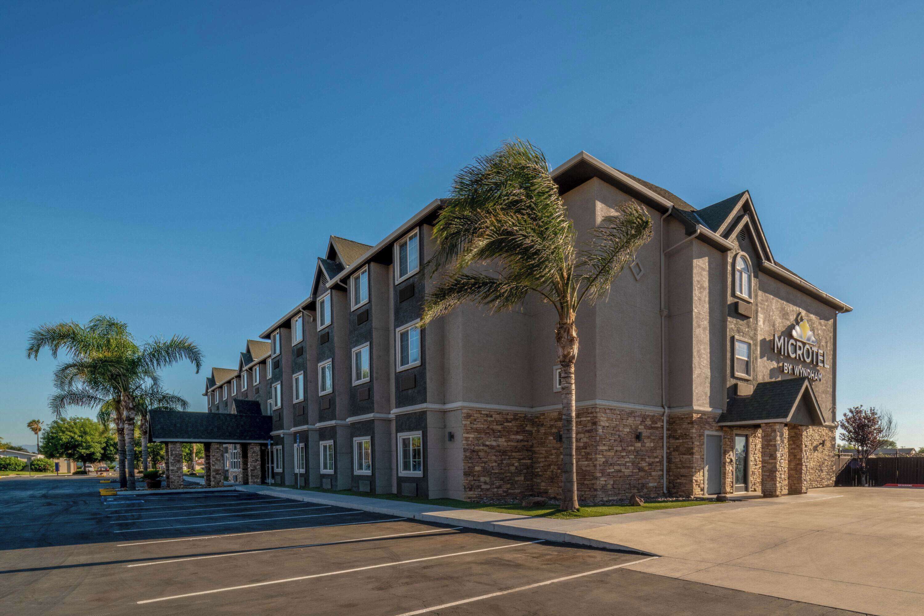 Un hotel de varios pisos con fachada de piedra, estacionamiento y una palmera frente a él.