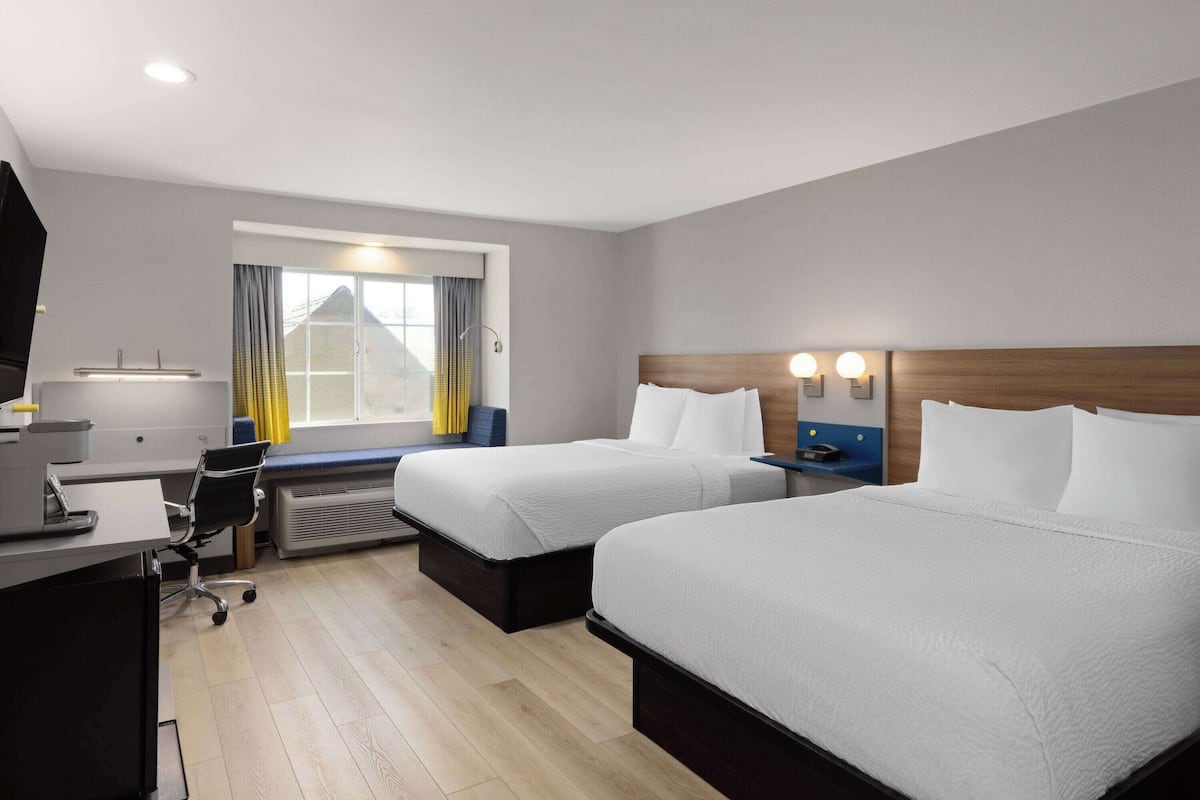A hotel room with two beds, a desk, a chair, a window with curtains, and a wall-mounted lamp.