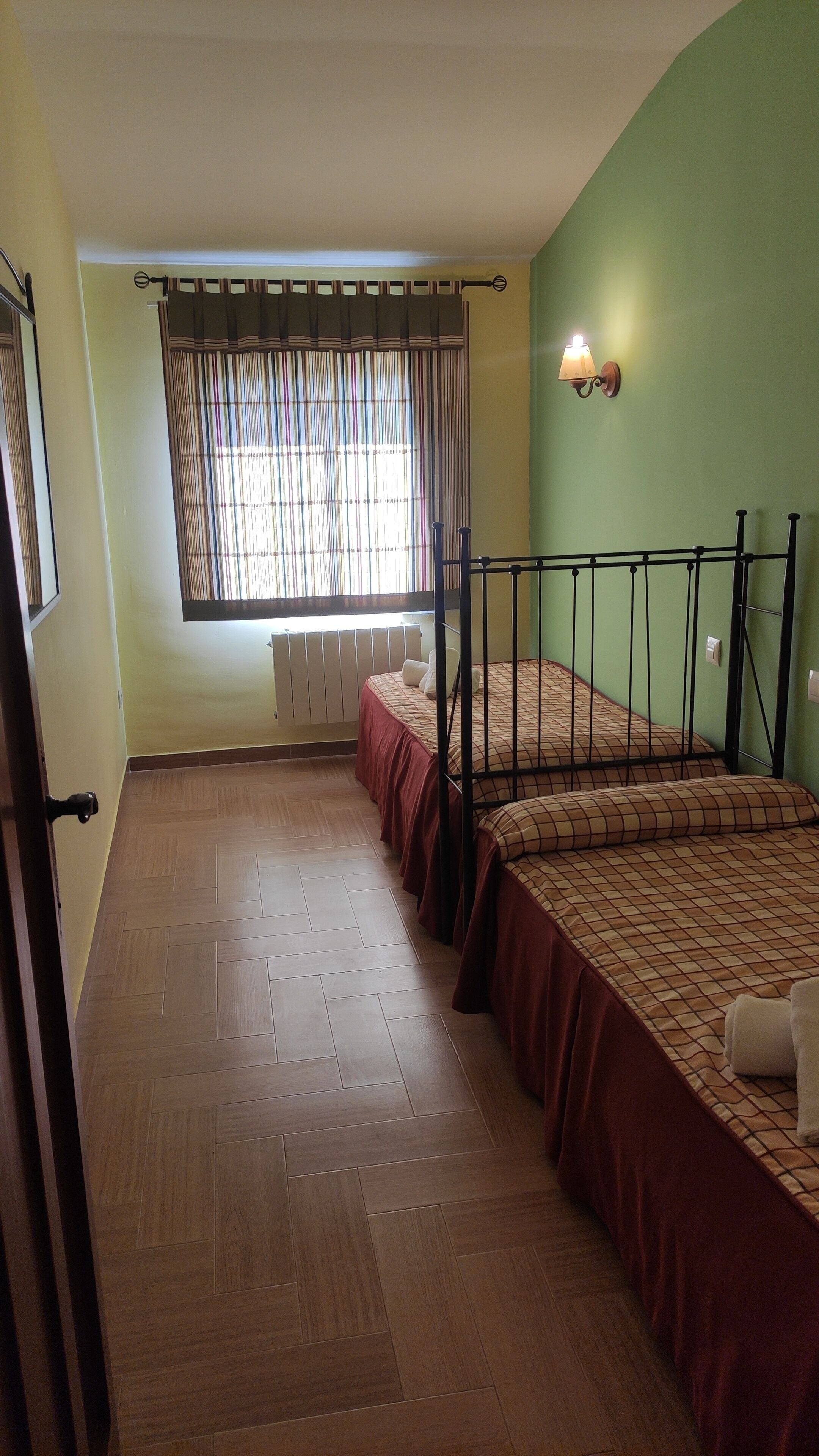 A room with two beds, a radiator, a window with curtains, and a wall-mounted light fixture.