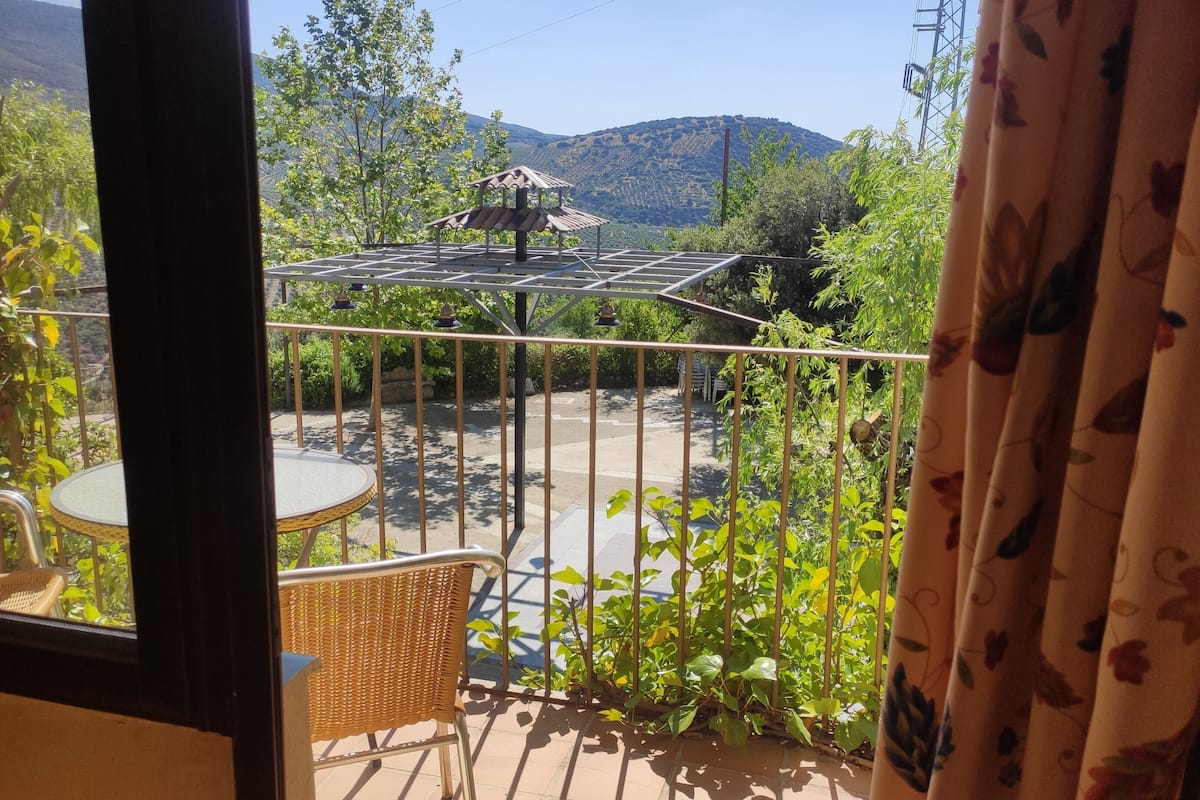 Un balcón con una mesa y sillas de mimbre, baranda metálica y vistas a un jardín y montañas.
