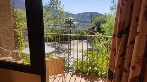 A balcony with a wicker table and chairs, a metal railing, and a view of a garden and mountains.
