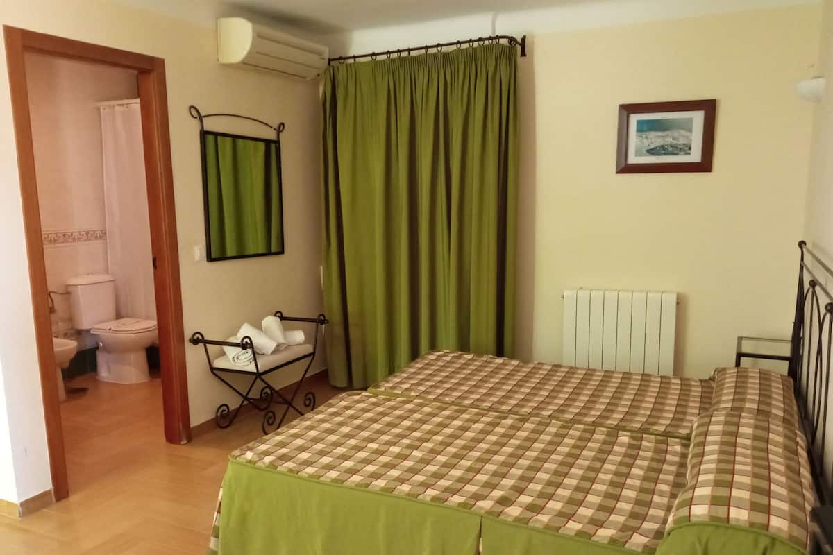 Un dormitorio con cama, un radiador, una cortina verde, un cuadro enmarcado y un baño visible a través de una puerta abierta.