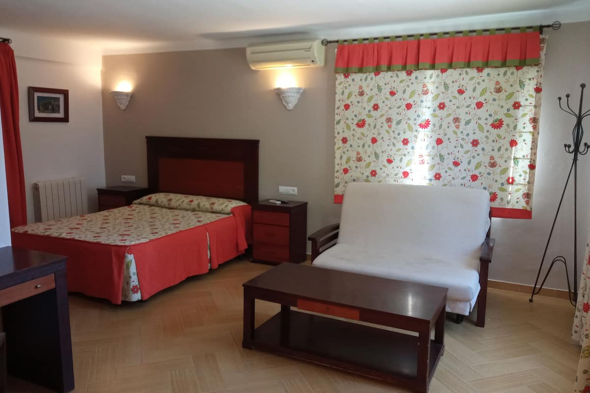 Habitación de hotel con cama, una silla, una mesita, una mesita de noche, un equipo de aire acondicionado y una ventana con cortinas florales.