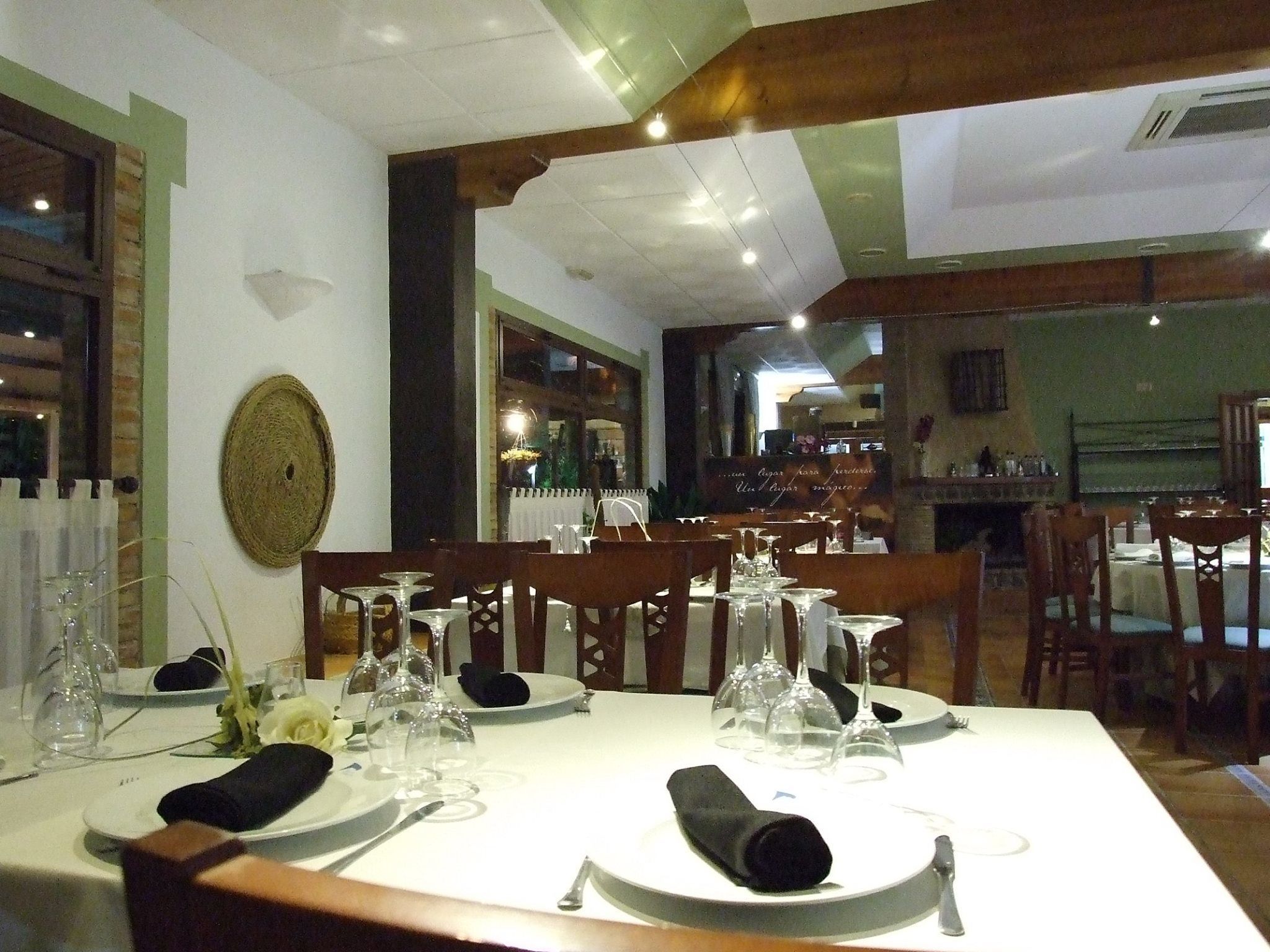 A restaurant with a long table set for dining, featuring a rustic wooden ceiling and a view of the kitchen in the background.