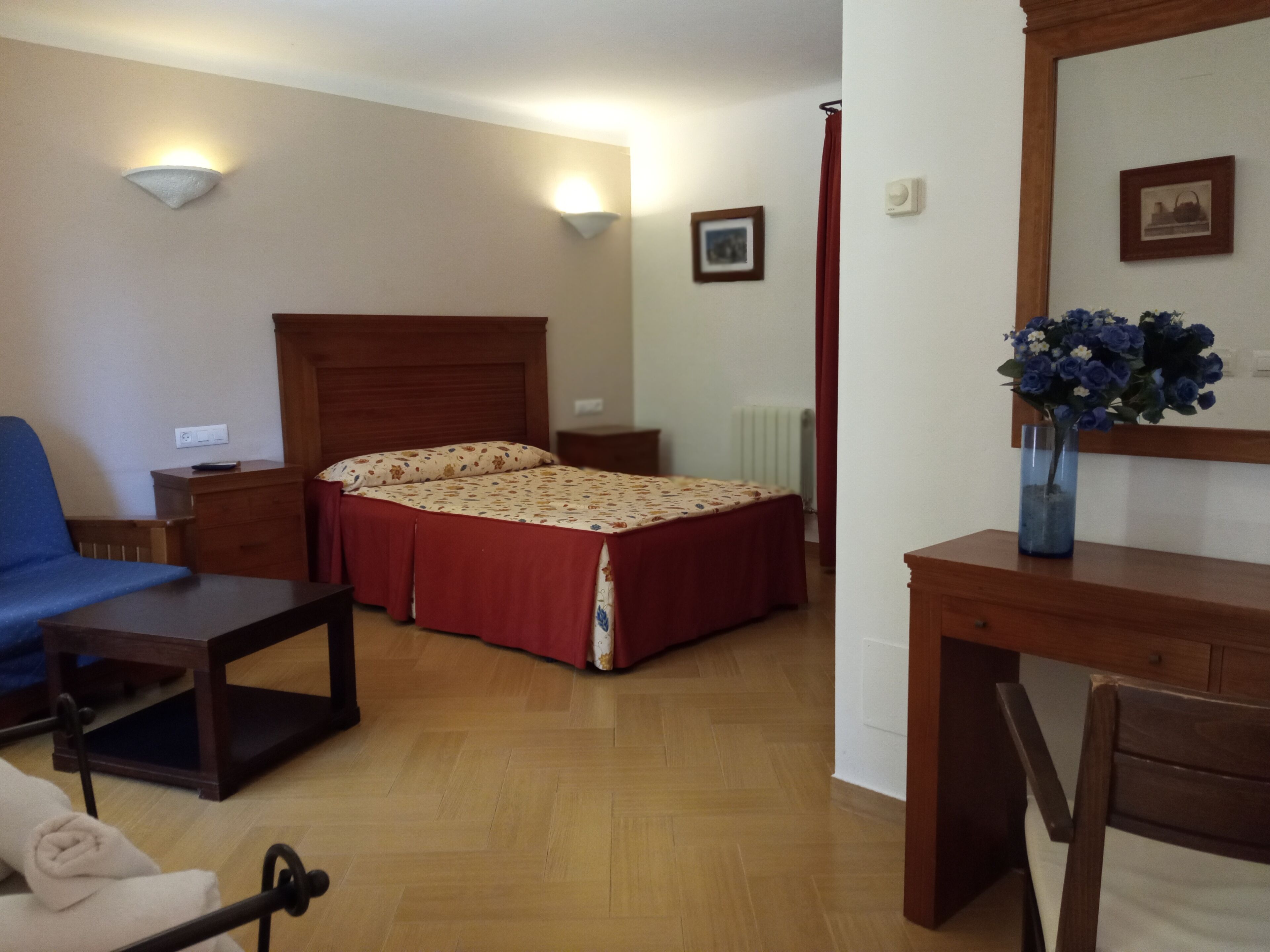 Habitación de hotel con cama, escritorio, silla, mesita de noche, lámpara y un florero con flores.