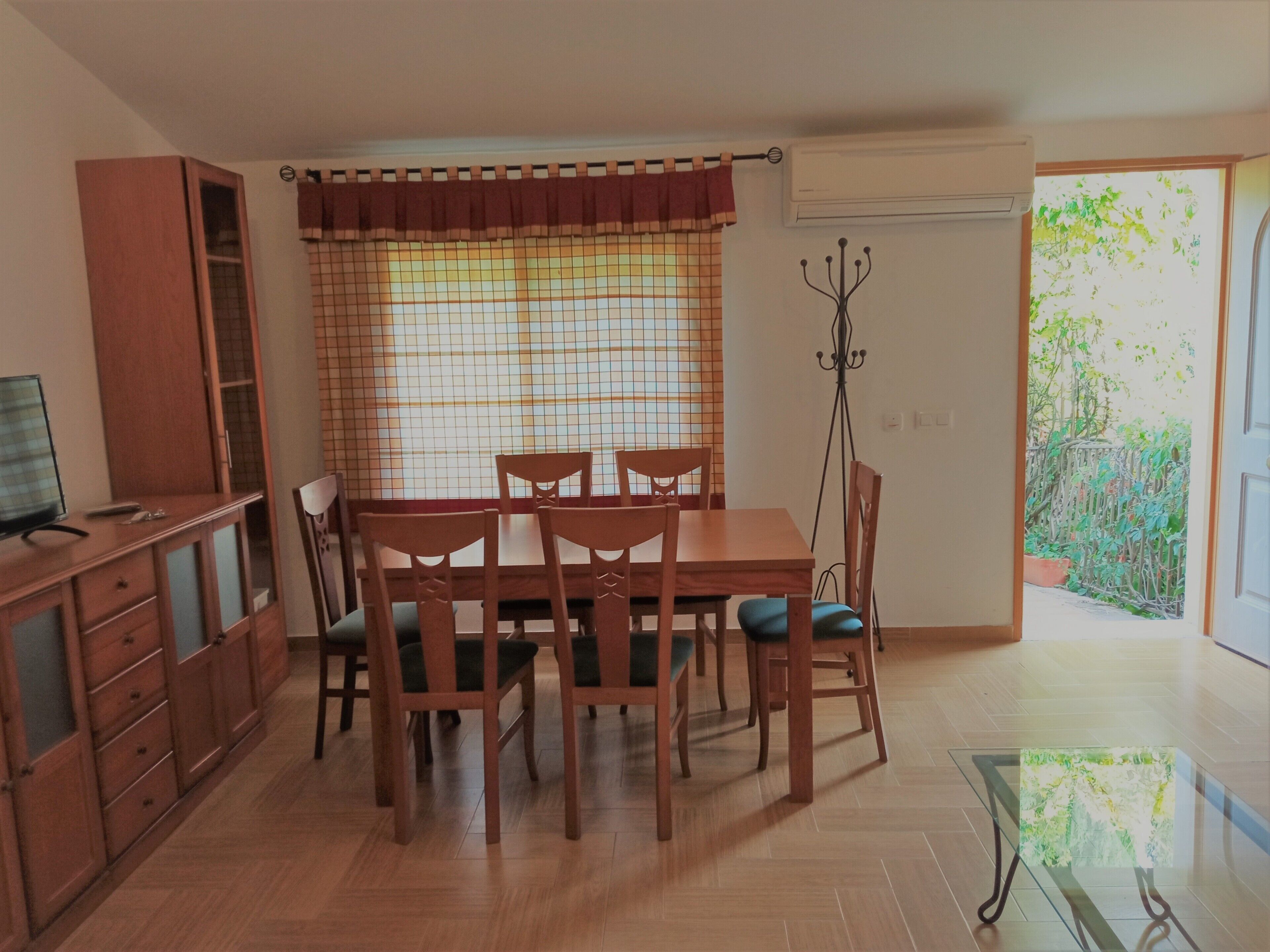 A dining area with a wooden table and chairs, a TV on a cabinet, and a window with a curtain.