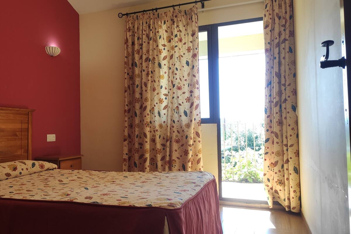 Un dormitorio con cama, cabecera de madera, ventana con cortinas y vista a zonas verdes.