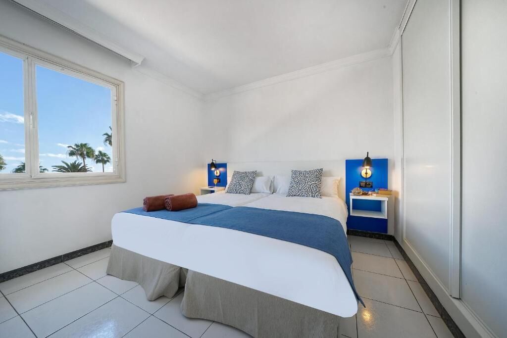 A hotel room with a bed, a nightstand, a lamp, and a window with a view of palm trees.