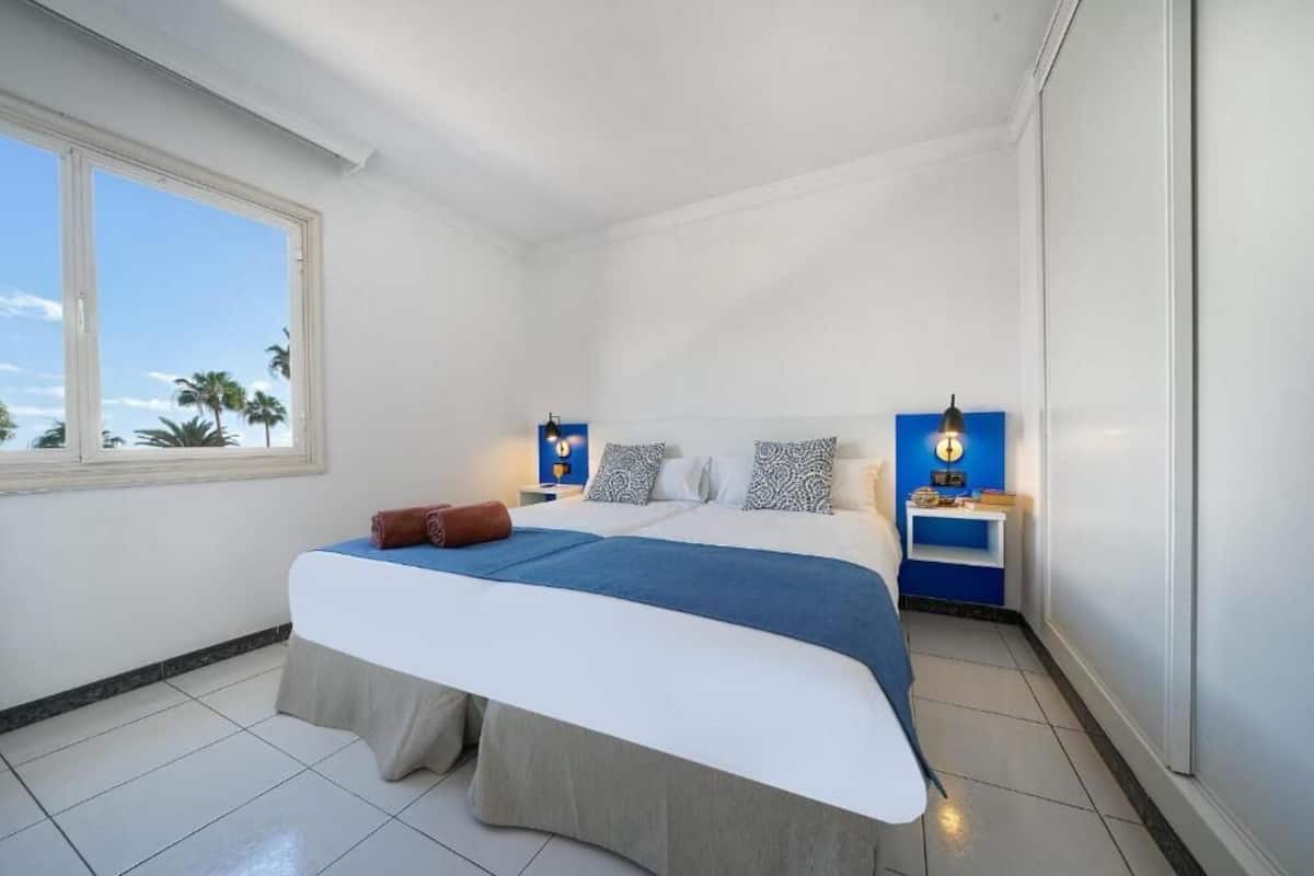A hotel room with a bed, a nightstand, a lamp, and a window with a view of palm trees.
