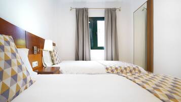 A hotel room with two beds, a wooden headboard, a bedside table with a lamp, and a window with curtains.
