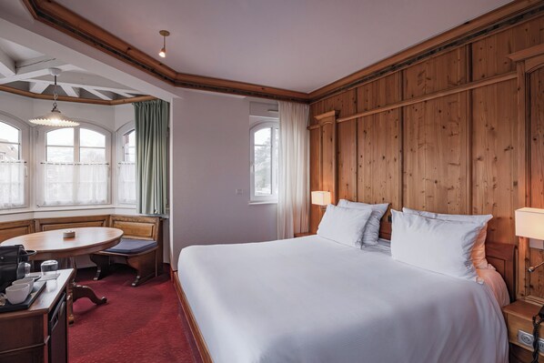 Una habitación de hotel con una cama grande, un cabecero de madera, una mesa de comedor con sillas y una ventana con cortinas.