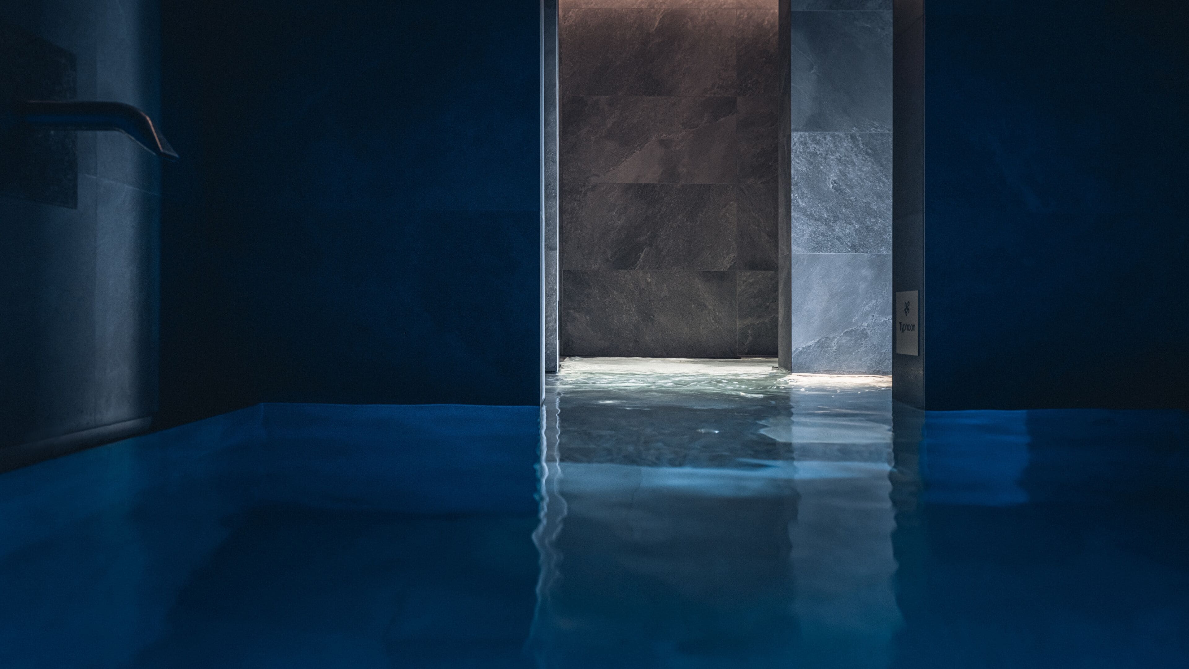 A modern indoor pool with a dark blue tiled floor and walls, a clear water surface, and a stone wall in the background.