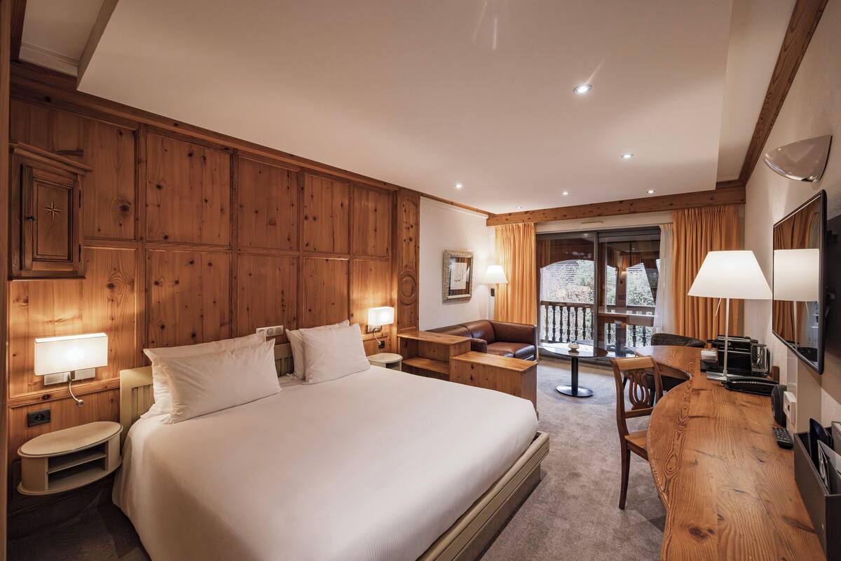 Une chambre d’hôtel avec un grand lit, des boiseries, un bureau avec une chaise, une petite table et un balcon avec des rideaux.