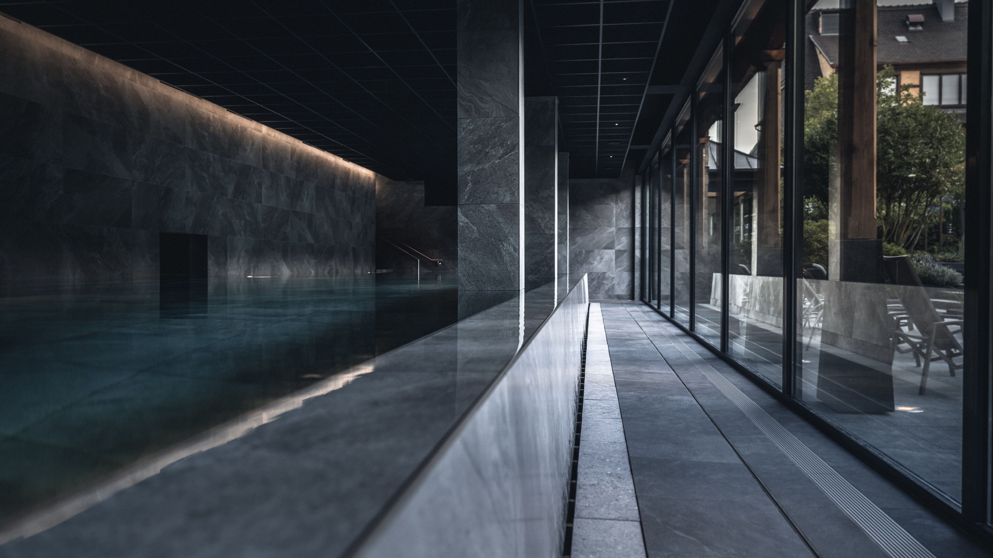 A modern indoor swimming pool with a minimalist design, featuring a long, narrow pool area and a glass wall on one side.