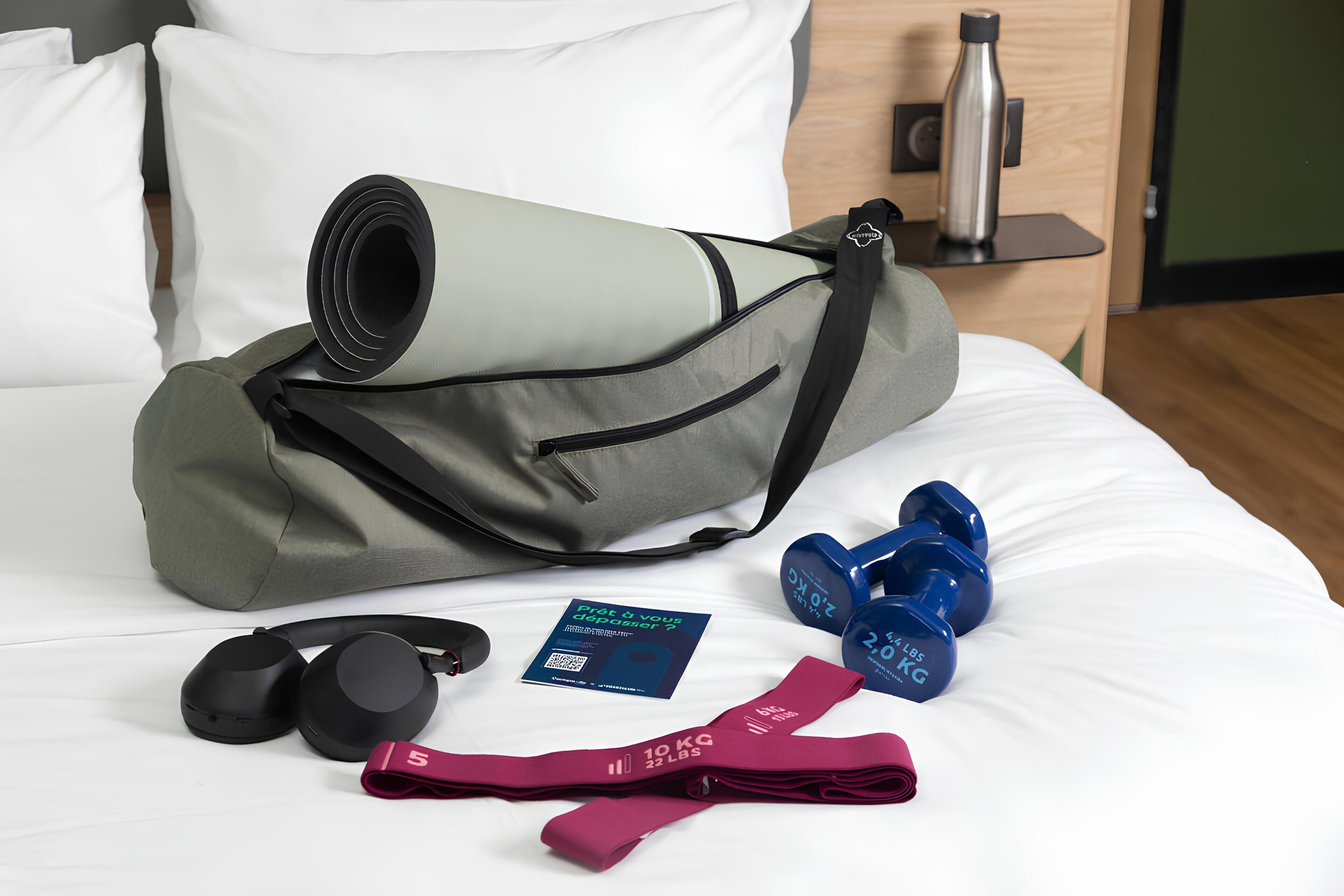 A neatly made bed with a yoga mat, dumbbells, resistance bands, and headphones on it.