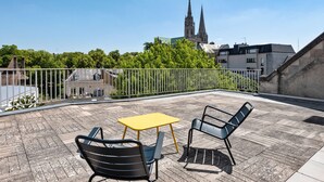 Una terraza en la azotea con una mesa amarilla y dos sillas, baranda metálica y una vista de una catedral al fondo.