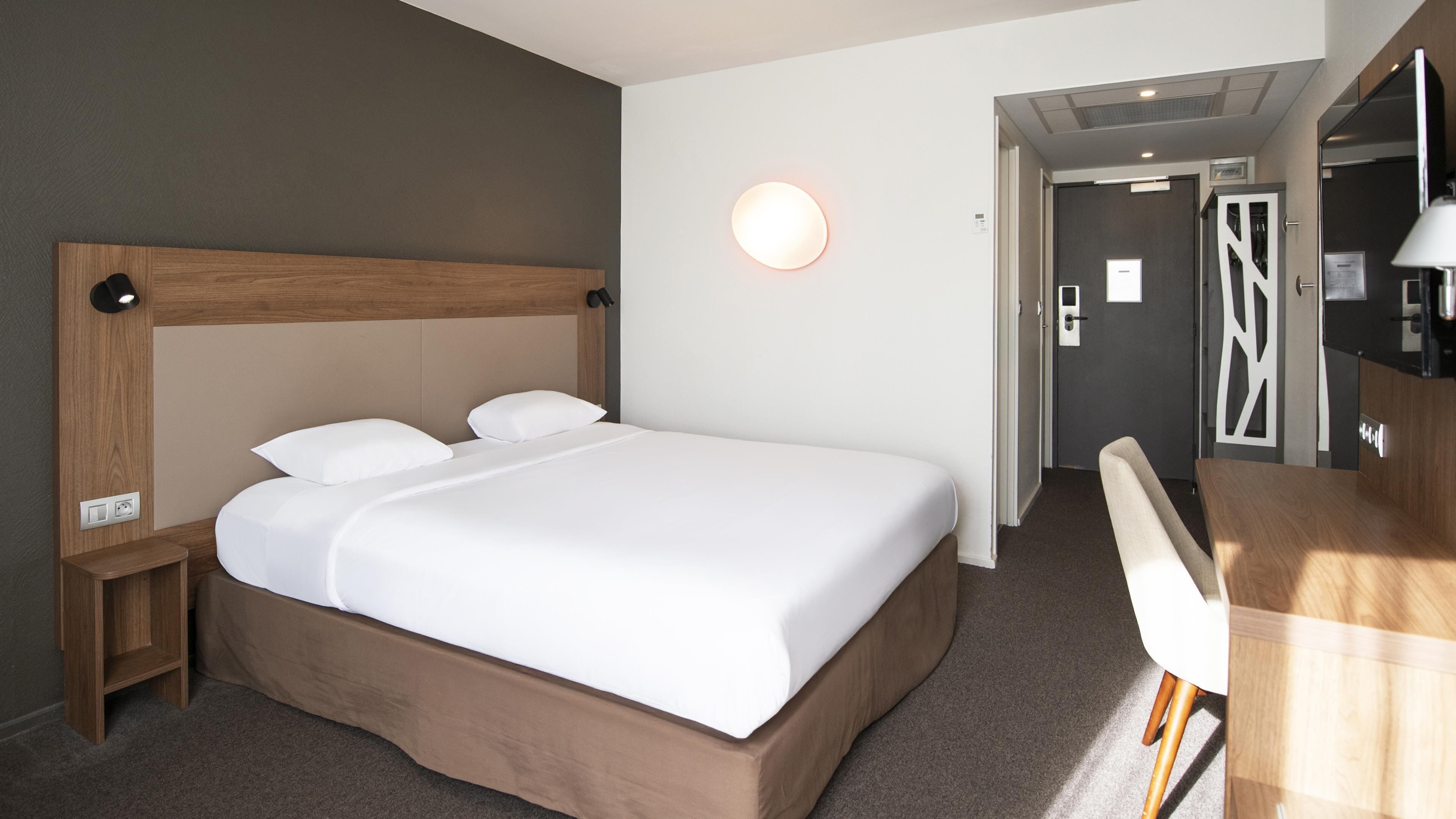 A hotel room with a large bed, a bedside table, a desk, and a chair.