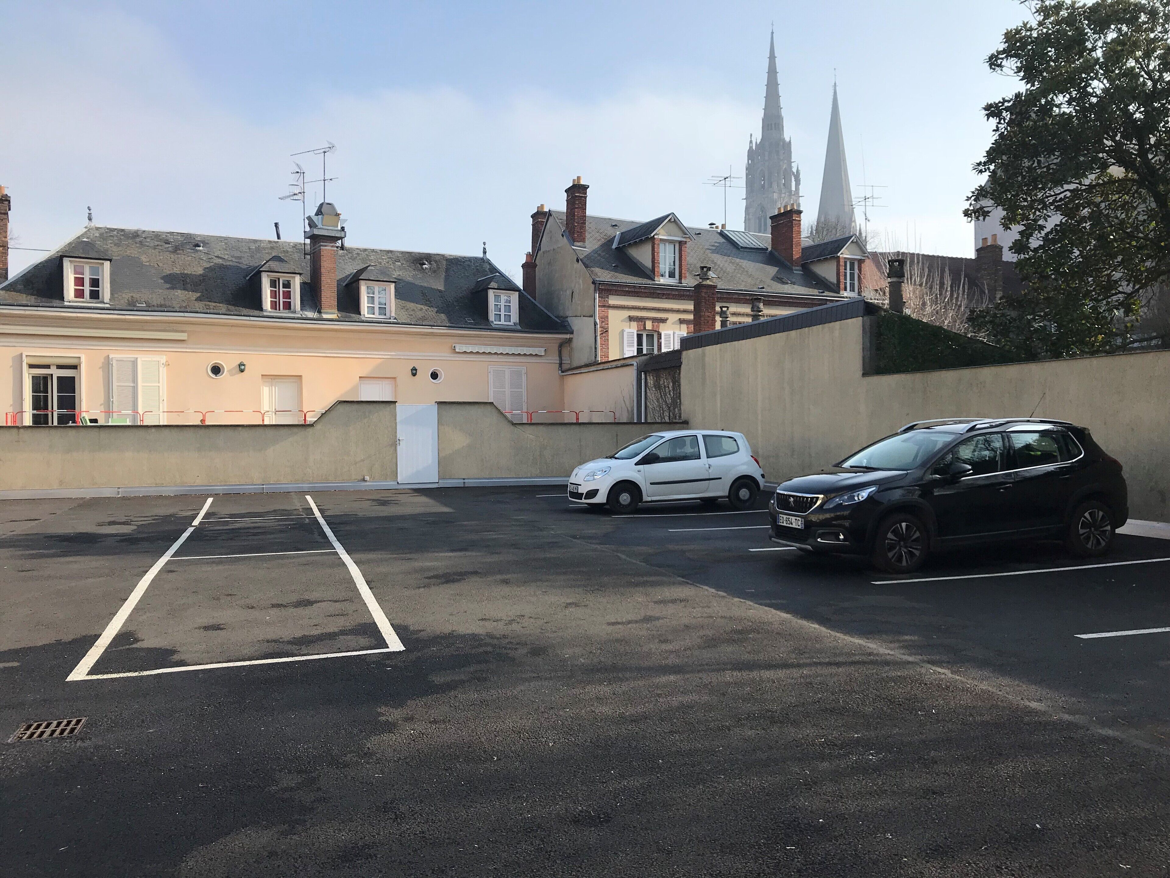 Een parkeerplaats met twee auto's, een gebouw met een beige gevel en een kerk met twee torens op de achtergrond.