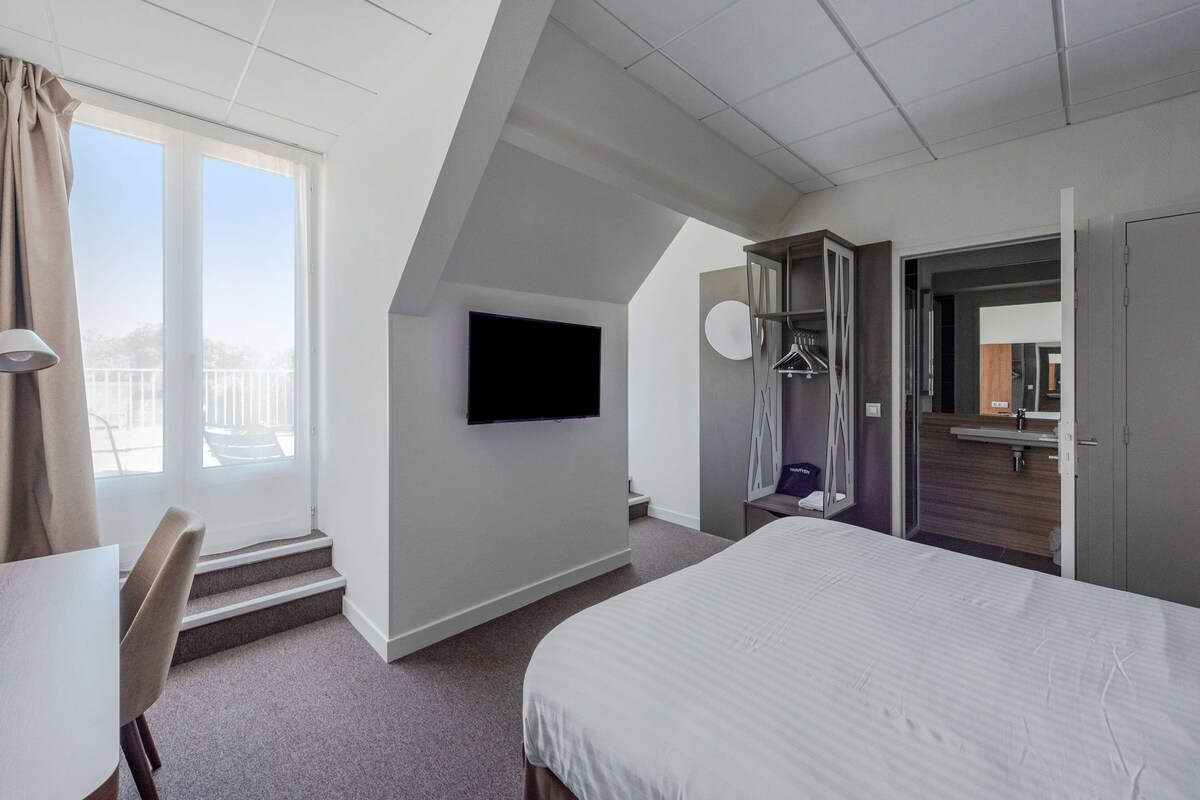 Una habitación de hotel moderna con una cama grande, un televisor de pantalla plana en la pared, un escritorio con una silla y vistas al exterior a través de una ventana.