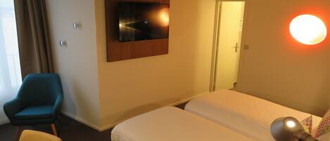 A hotel room with two beds, a TV mounted on the wall, a desk with a chair, and a lamp.