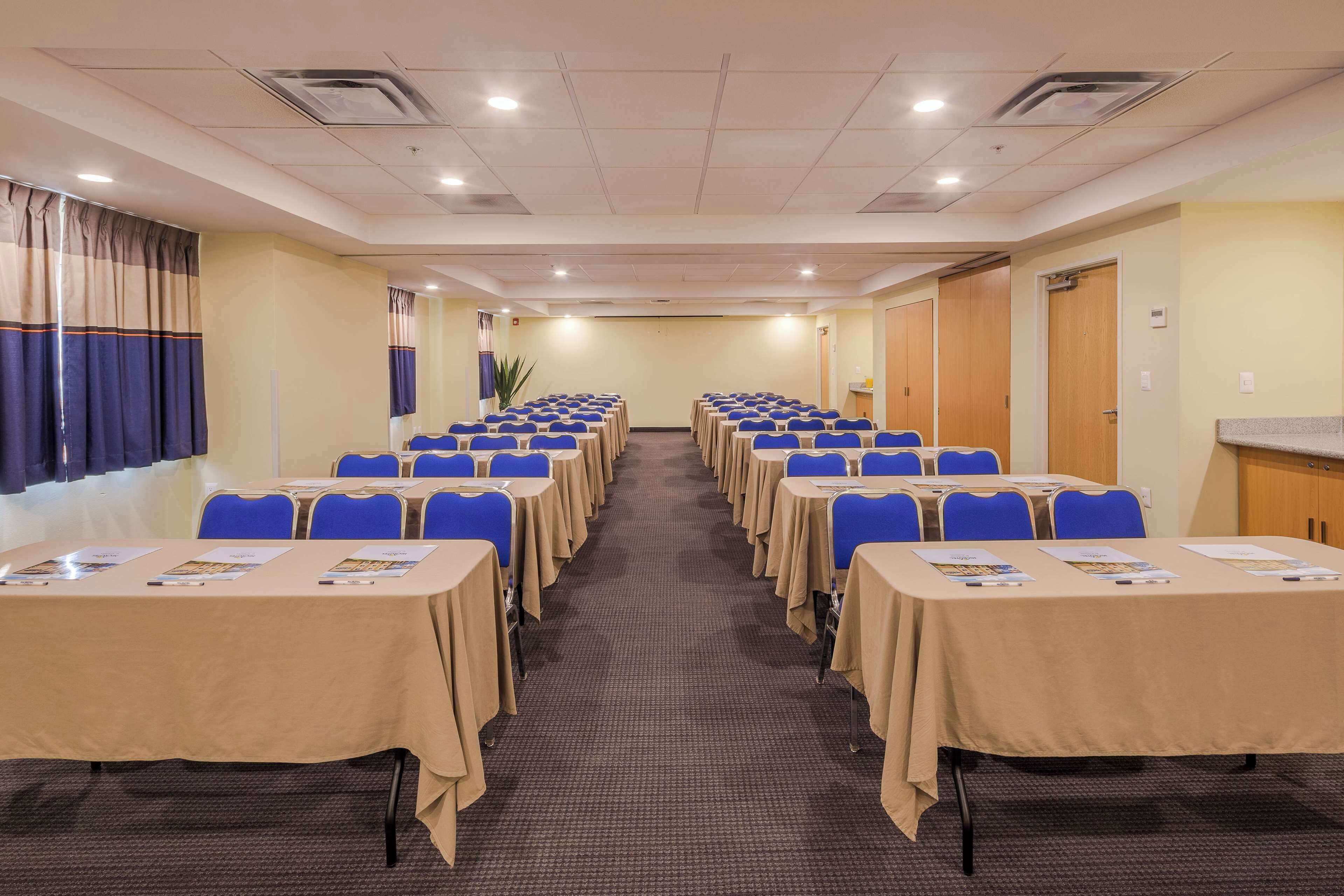 Un salón de conferencias con filas de mesas y sillas, suelo alfombrado y una pared sencilla con una puerta y una planta.