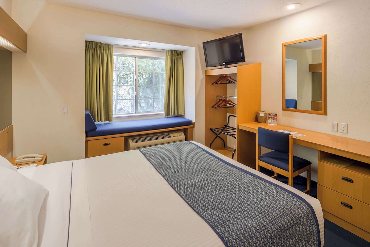 A hotel room with a bed, a desk with a chair, a television, a mirror, and a window with curtains.