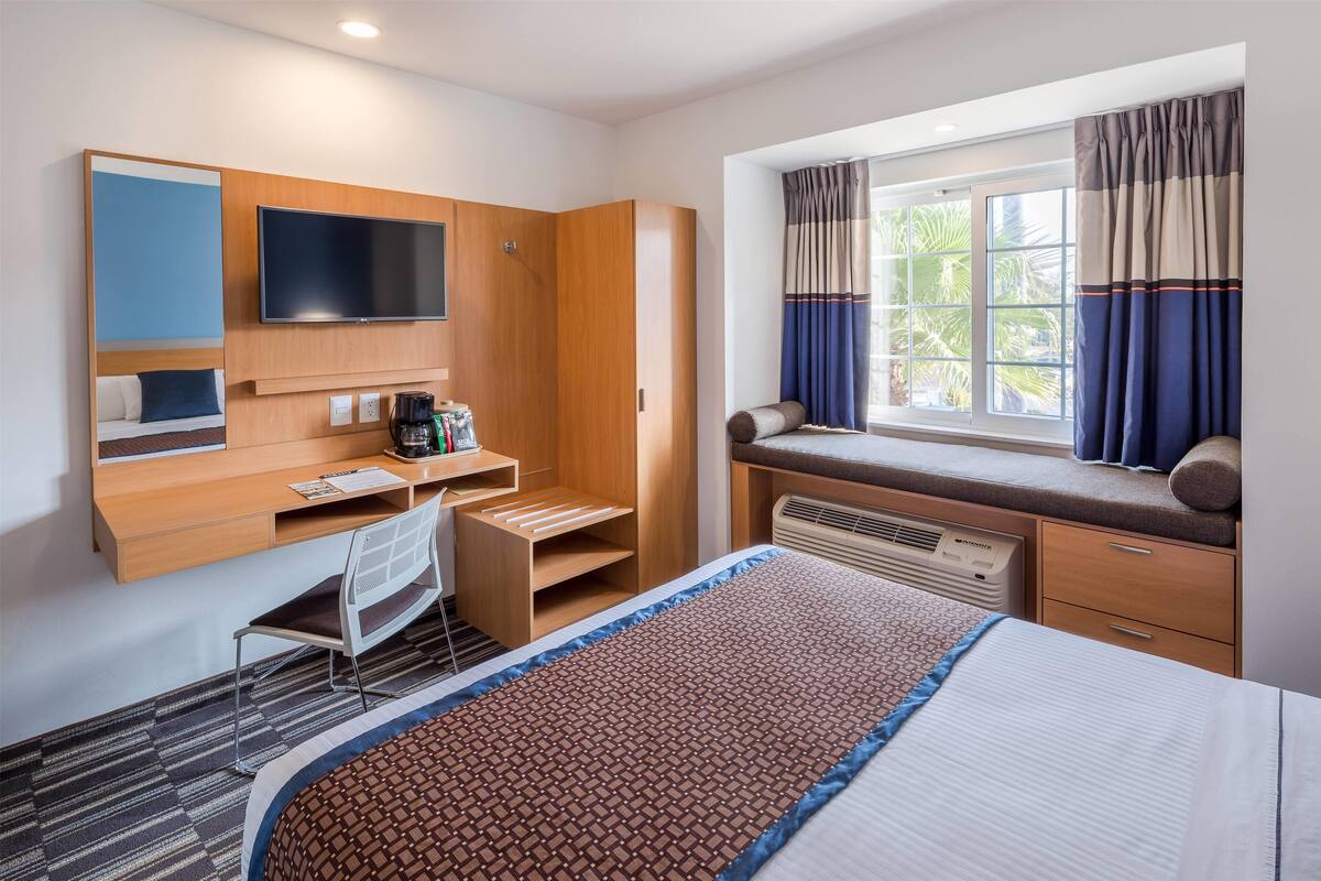 A hotel room with a bed, a desk with a chair, a TV, a window with curtains, and a built-in bench with a view.